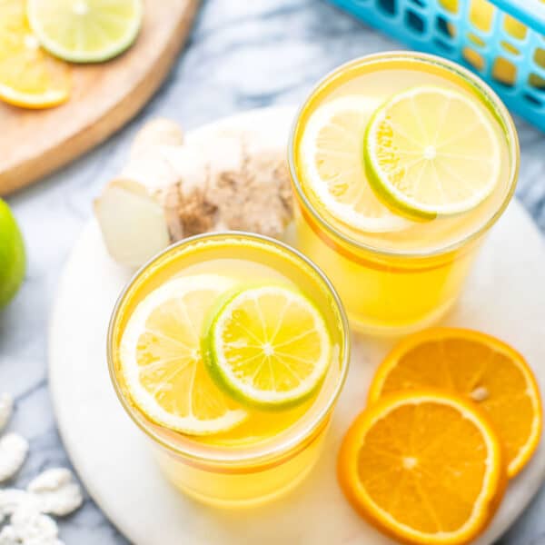 a couple of glasses of Homemade Electrolyte Drink garnished with lemon, lime and orange slices in front of a basket of citrus fruit and a cutting board with fruit slices