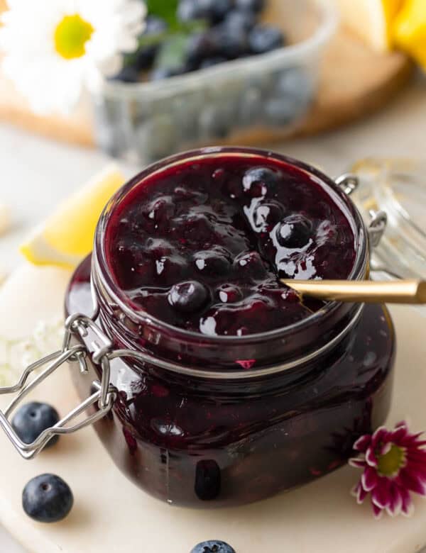 homemade blueberry filling for cakes and pies in a jar with a spoon surrounded by fresh berries