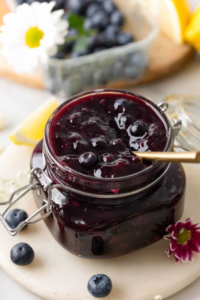 homemade blueberry pie filling for cakes and pies in a jar with a spoon surrounded by fresh berries