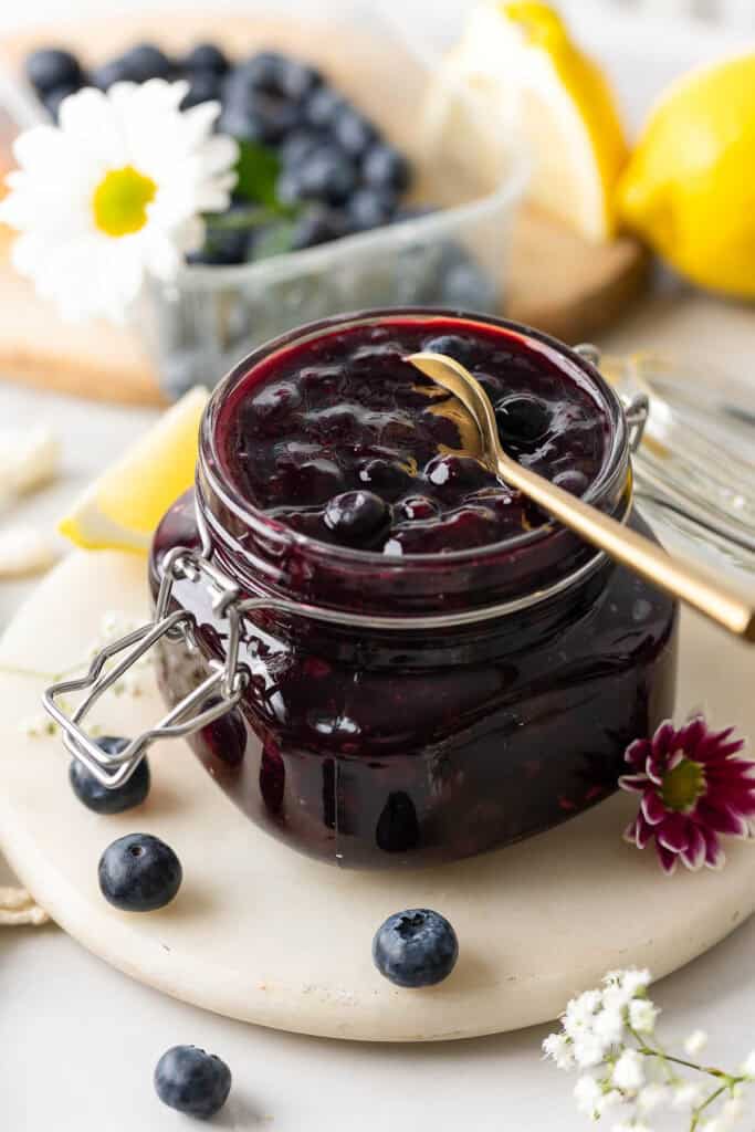 homemade blueberry pie filling for cakes and pies in a jar with a spoon surrounded by fresh berries, flowers and lemons