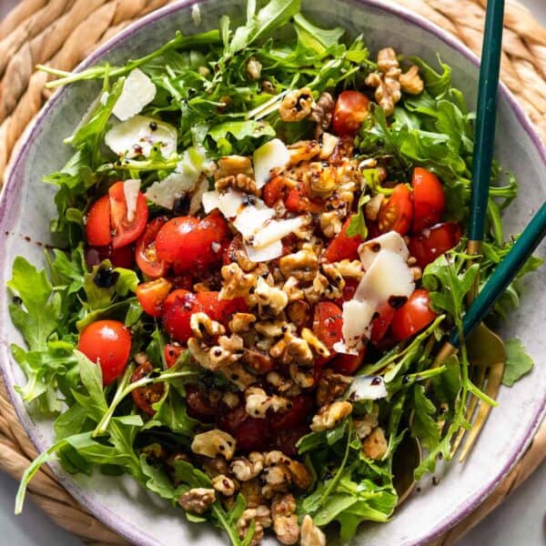 a Tomato Arugula Salad with toasted walnuts and shaved parmesan drizzled with olive oil and a balsamic vinegar reduction in a salad bowl with serving spoons
