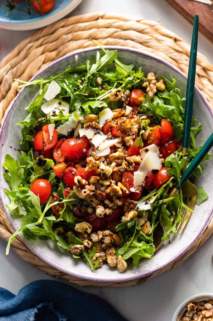 a Tomato Arugula Salad with toasted walnuts and shaved parmesan drizzled with olive oil and a balsamic vinegar reduction in a salad bowl with serving spoons