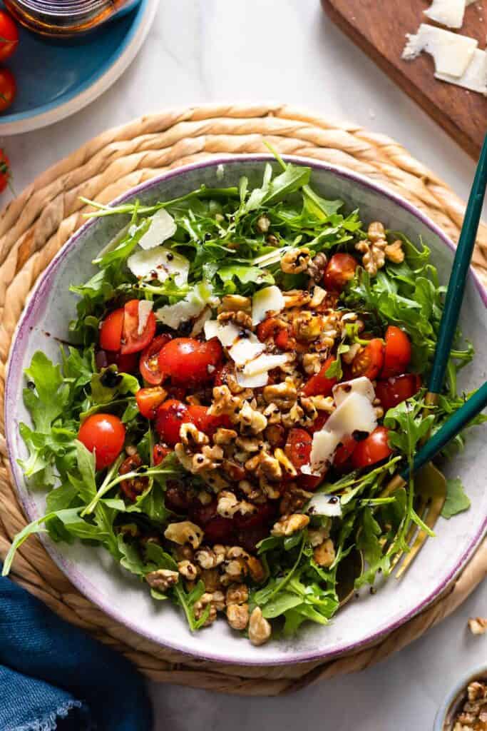 a Tomato Arugula Salad with toasted  walnuts and shaved parmesan drizzled with olive oil and a balsamic vinegar reduction in a salad bowl with serving spoons