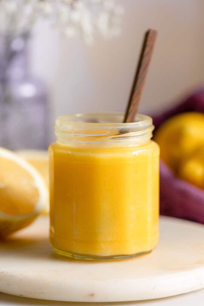 homemade lemon curd in a small jar with a spoon inside and fresh lemons surrounding