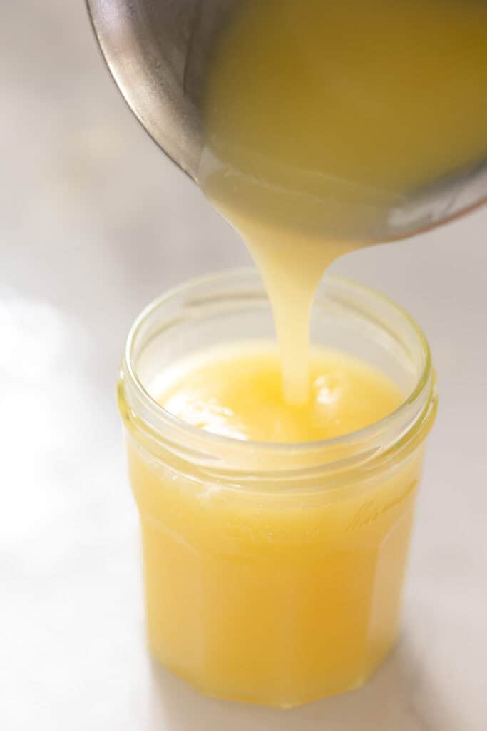 homemade vegan lemon curd pouring out of a sauce pot and into a storage jar