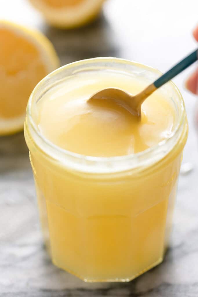 Homemade Vegan Lemon Curd in a jar stirred with a spoon with lemons in the back