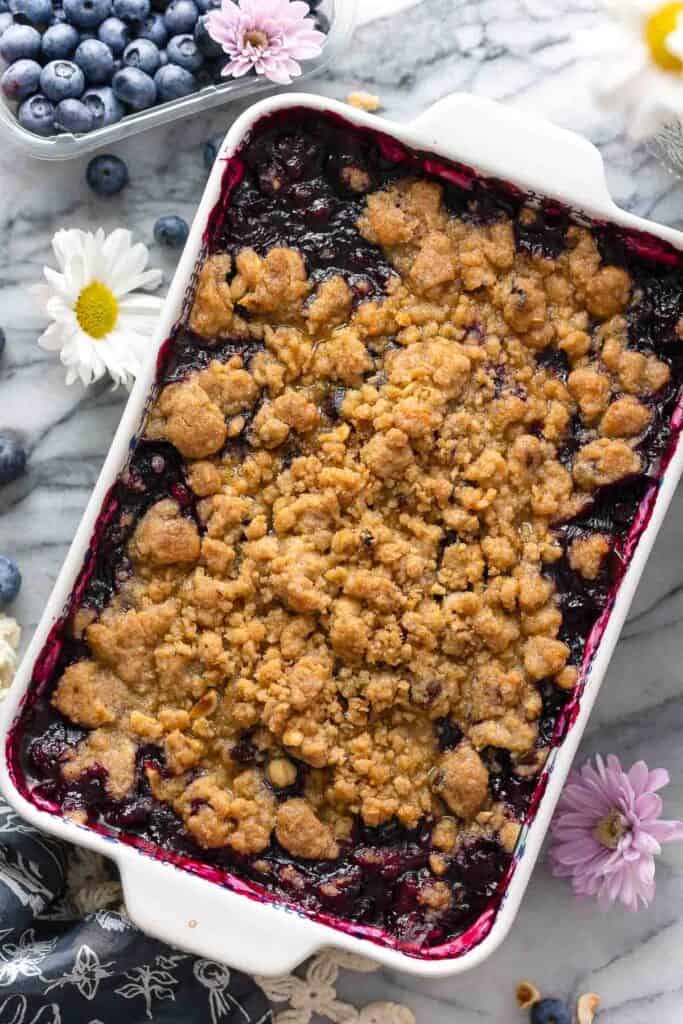 Gluten Free Blueberry Crumble in a baking dish surrounded by fresh blueberries and flowers