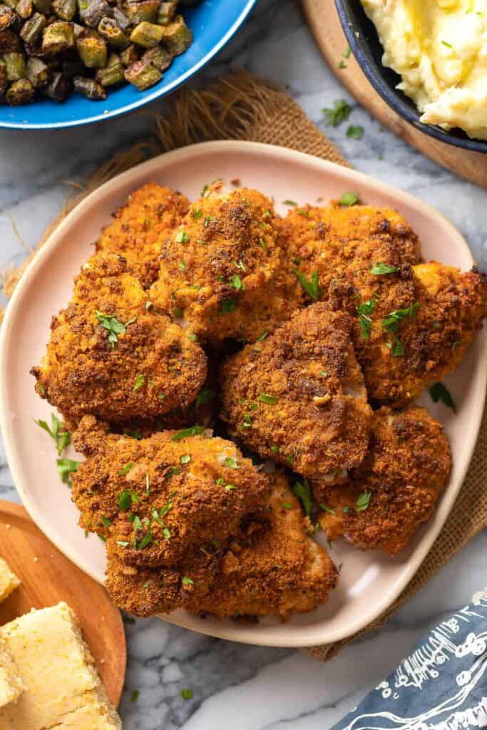 Oven Fried Chicken Thighs on a plate with fresh parsley on top with roasted okra, mashed potatoes and cornbread on the side