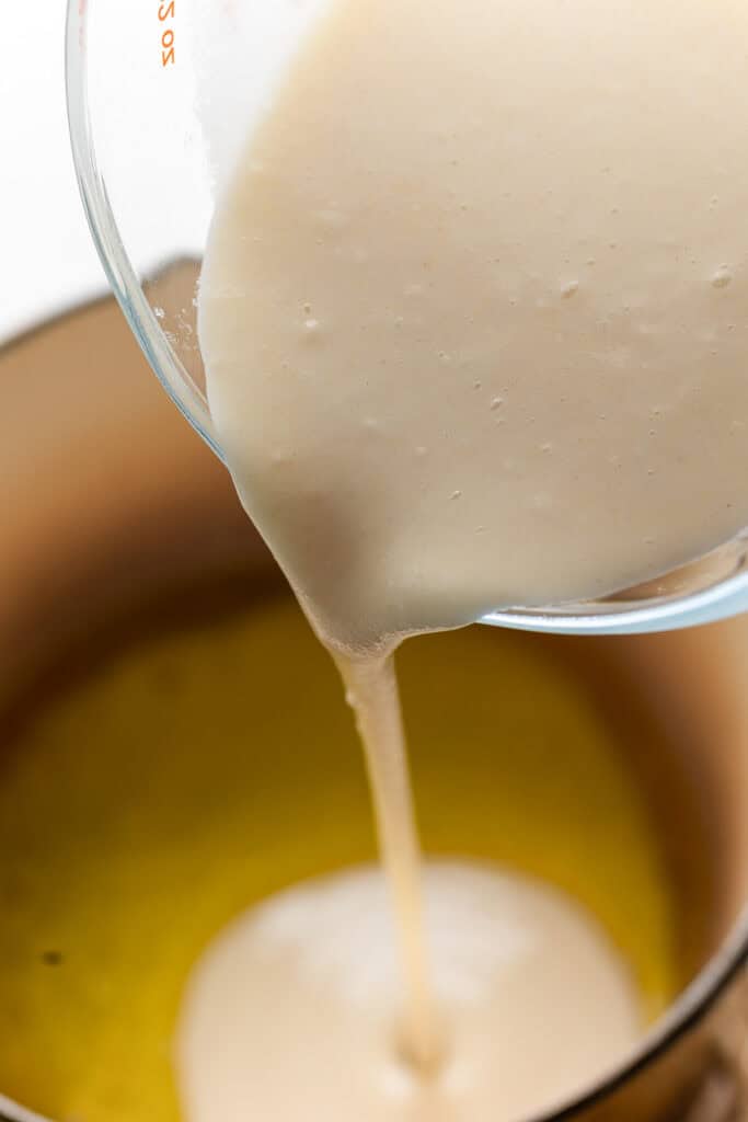 a water and flour mixture pouring from a measuring jug into a pot of melted butter with text: 'add flour mixture to butter'