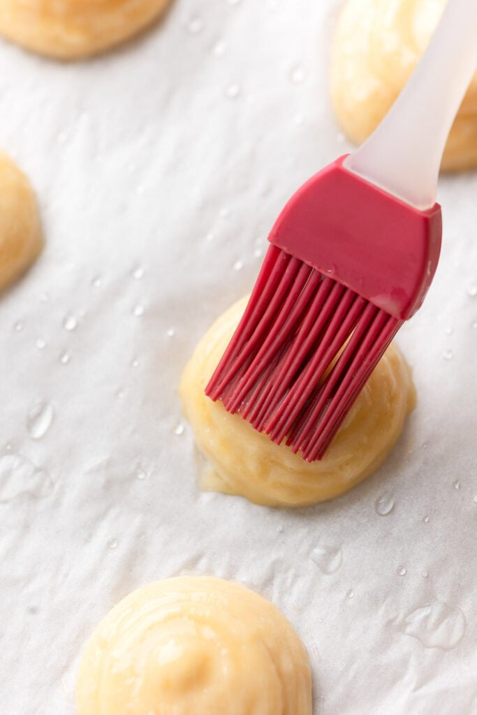 a pastry brush brushed whisked egg on top of Gluten free choux batter piped on a tray 