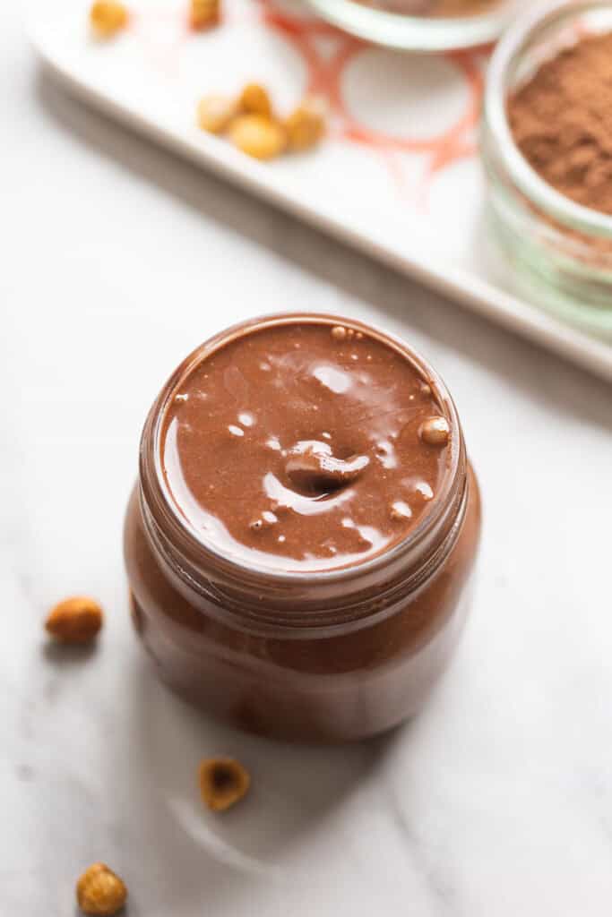 a jar full of homemade Nutella (chocolate hazelnut spread) with loose hazelnuts in the back