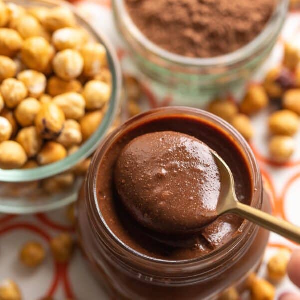 homemade nutella in a jar with spoon inside and toasted hazelnuts and cocoa powder in the back