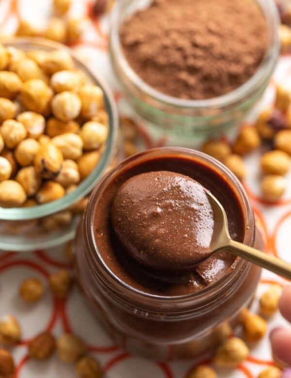 homemade nutella in a jar with spoon inside and toasted hazelnuts and cocoa powder in the back