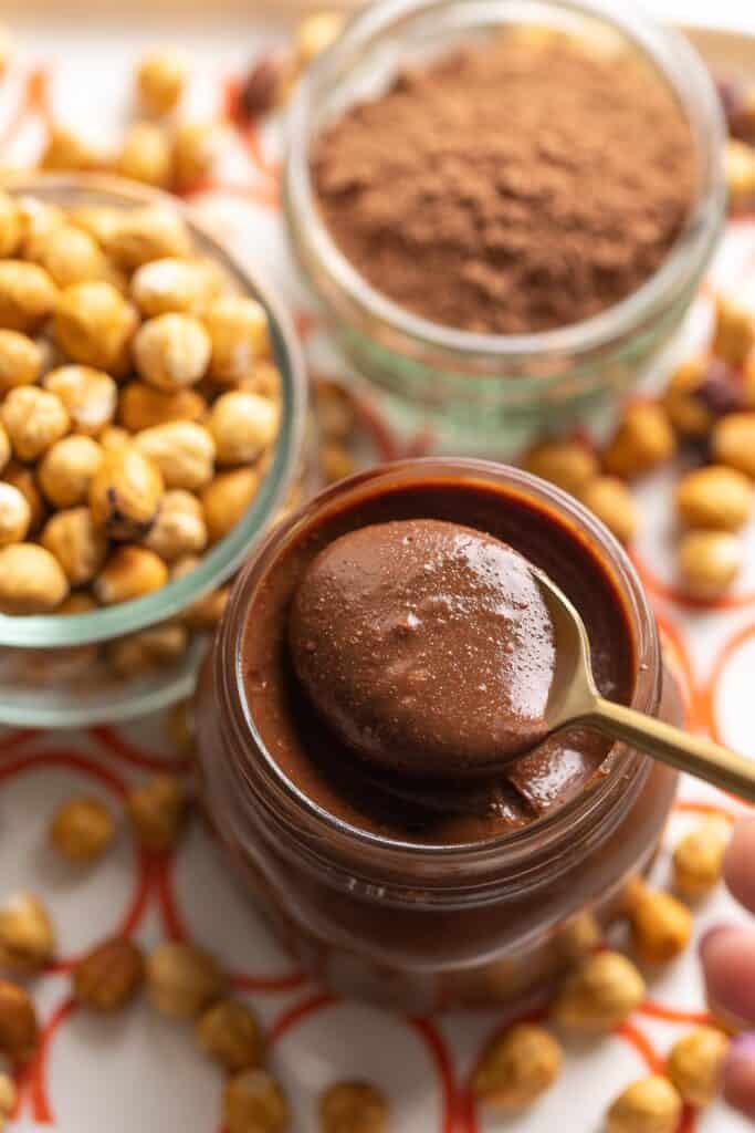 a spoonful of homemade nutella in a jar with roasted hazelnuts and cocoa powder in the back