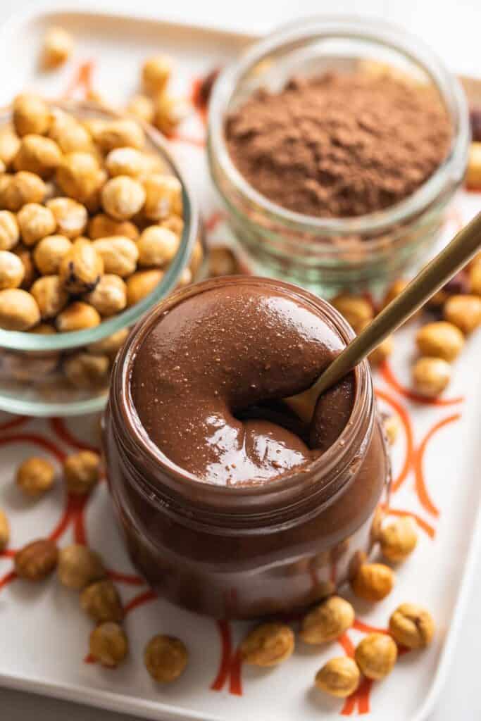 homemade nutella in a jar with  spoon inside and toasted hazelnuts and cocoa powder in the back