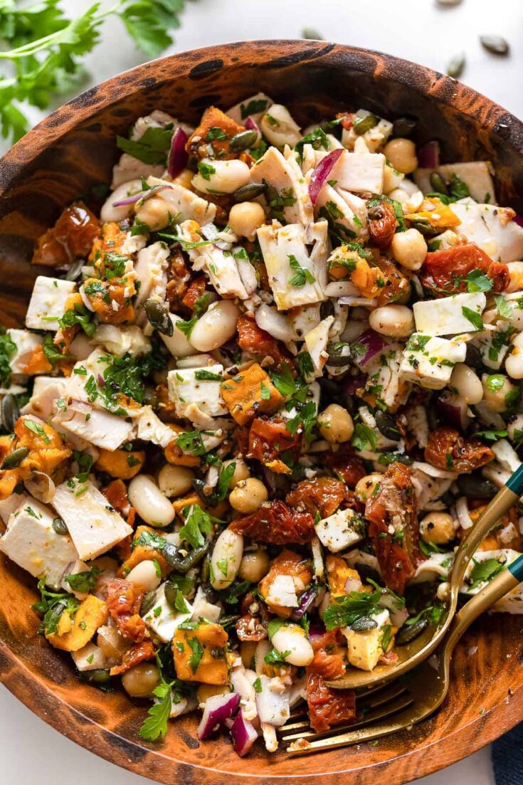 a Mediterranean Dense Bean Salad in a large salad bowl full of cannellini beans, chickpeas, sun-dried tomatoes, fresh parsley, roasted sweet potatoes, chopped feta and sliced deli turkey breast tossed in a lemon vinaigrette