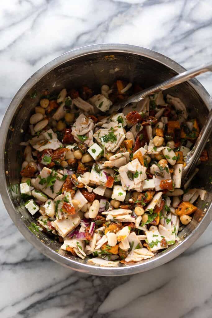 a mediterranean dense bean salad made up of white beans, fresh parsley, turkey slices, feta, sun-dried tomato and sweet potato in a large salad bowl dressed in a lemon vinaigrette 