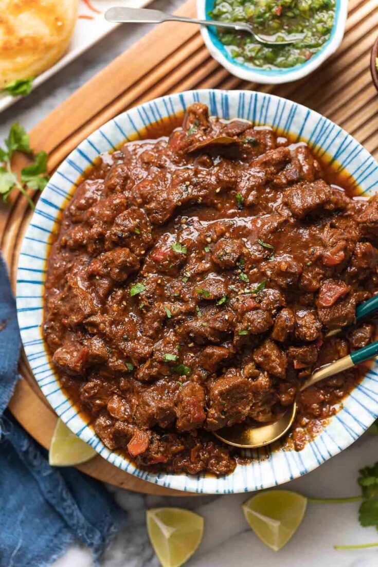 a serving bowl full of extra Saucy Carne Desmechada topped with chopped cilantro and fresh black pepper on a serving board with lime wedges on the sides