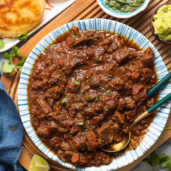 a serving bowl full of extra Saucy Carne Desmechada topped with chopped cilantro and fresh black pepper on a serving board with lime wedges and arepas on the sides