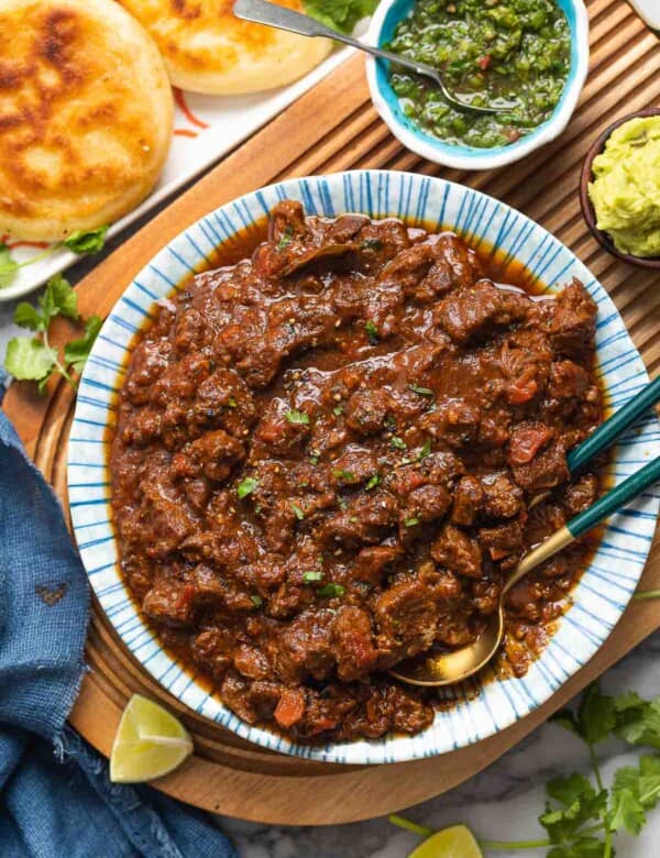 a serving bowl full of extra Saucy Carne Desmechada topped with chopped cilantro and fresh black pepper on a serving board with lime wedges and arepas on the sides