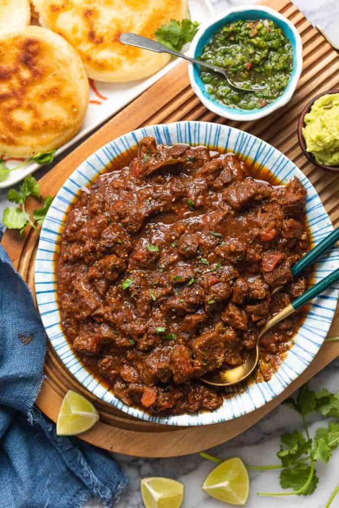 a serving bowl full of extra Saucy Carne Desmechada topped with chopped cilantro and fresh black pepper on a serving board with lime wedges and arepas on the sides 