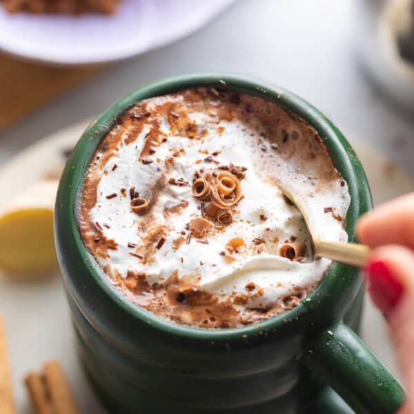 a mug of chai hot chocolate with whipped cream and cinnamon on top stirred with a spoon