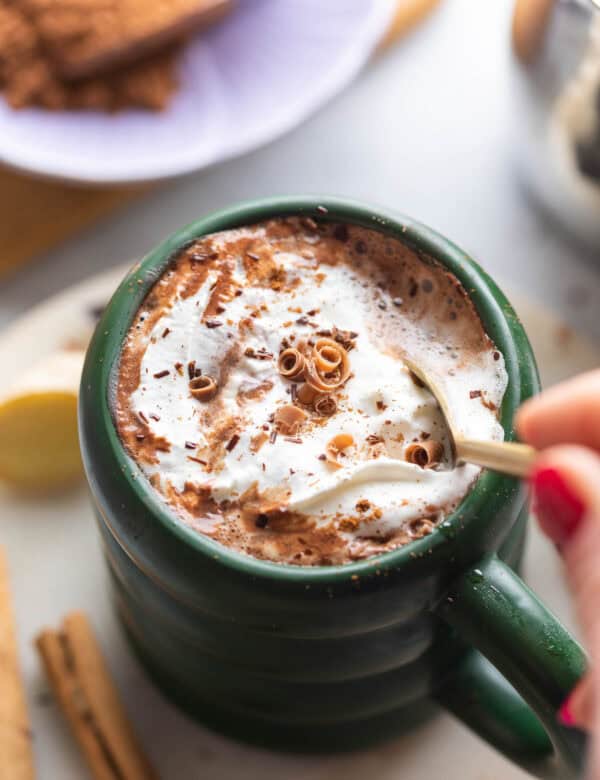 a mug of chai hot chocolate with whipped cream and cinnamon on top stirred with a spoon