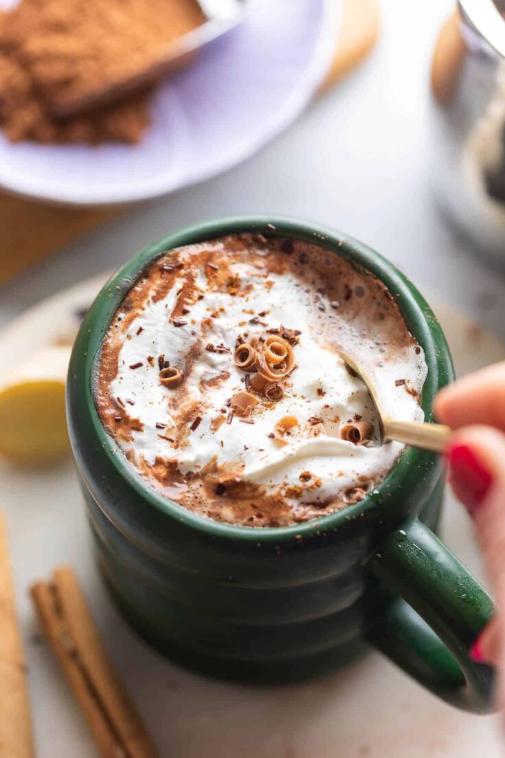 a mug of chai hot chocolate with whipped cream and cinnamon on top stirred with a spoon