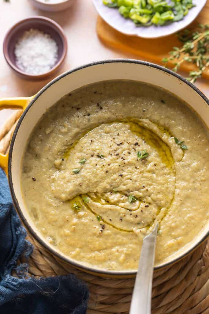 Roasted Cauliflower and Garlic Soup in a soup pot with a ladle topped with an olive oil dried and fresh thyme leaves with a bowl of salt on the side