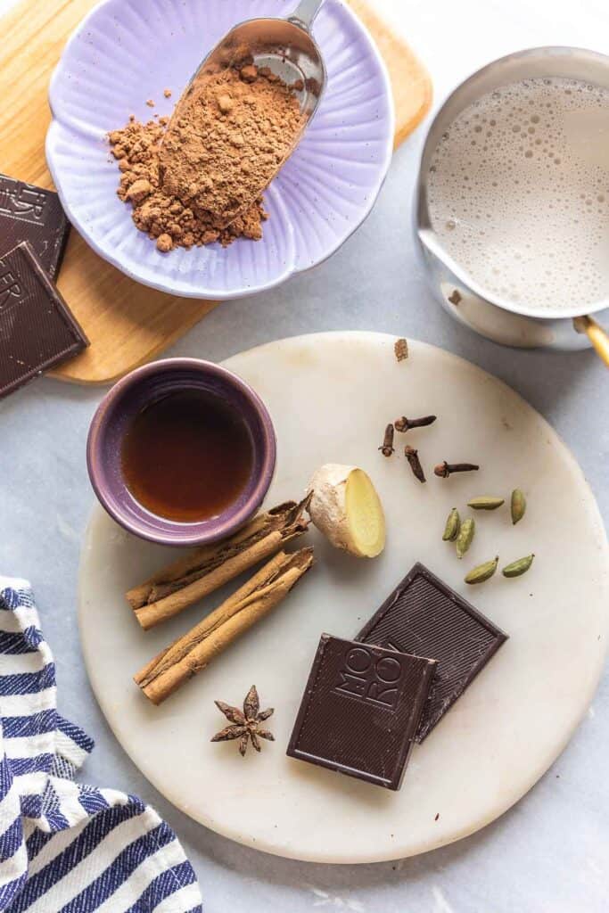 the whole ingredients needed to make homemade spices chai hot chocolate: cinnamon sticks, cardamom pods, cloves, fresh ginger, star anise, chocolate, cocoa powder and milk