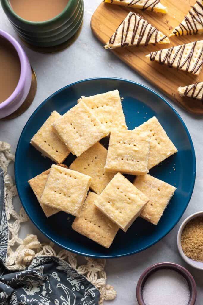 Plain gluten free shortbread piled on a plate. In the back there are shortbread cookies cut into triangles and drizzled with chocolate