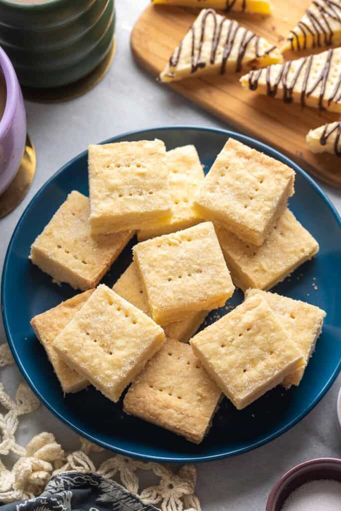 Gluten Free Shortbread cut into squares on a plate with some shortbread cookies cut into triangles and drizzled with chocolate on a wooden cutting board in the back