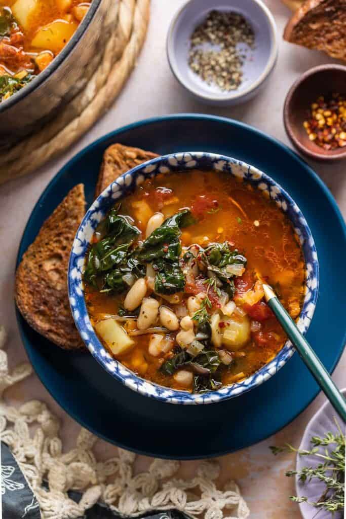 A bowl full of Bacon Kale Bean Soup on a plate with a couple slices of gluten free toast; chili flakes and black pepper in small bowls on the side