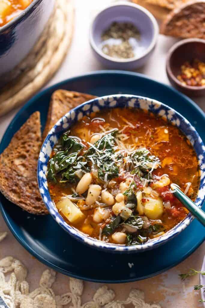 A bowl full of Bacon Kale Bean Soup topped with grated parmesan on a plate with a couple slices of gluten free toast