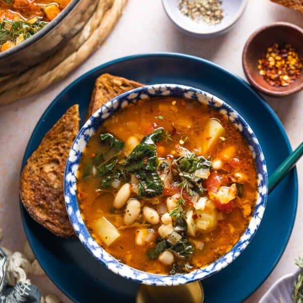 A bowl full of Bacon Kale Bean Soup spoon on a plate with a couple slices of gluten free toast and a spoon; chili flakes and black pepper in small bowls on the side