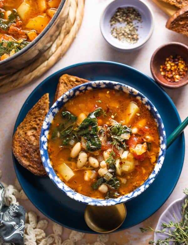 A bowl full of Bacon Kale Bean Soup spoon on a plate with a couple slices of gluten free toast and a spoon; chili flakes and black pepper in small bowls on the side