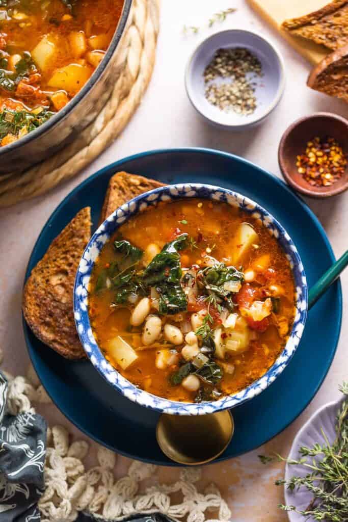 A bowl full of Bacon Kale Bean Soup on a plate with a couple slices of gluten free toast and a spoon; chili flakes and black pepper in small bowls on the side