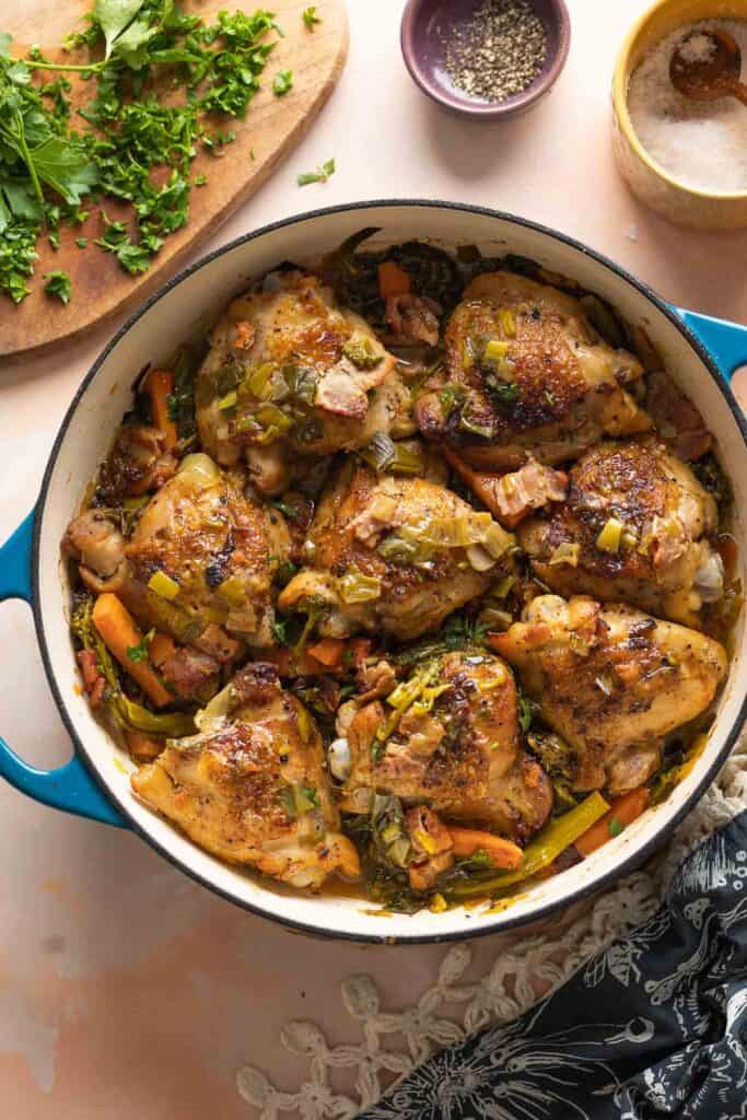 A large casserole dish full of White Wine Braised Chicken Thighs and vegetables with fresh parsley, salt and pepper on the side.