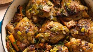 A ceramic casserole dish full of white wine braised chicken thighs and assorted vegetables including carrots and broccoli with fresh parsley chopped on the side.