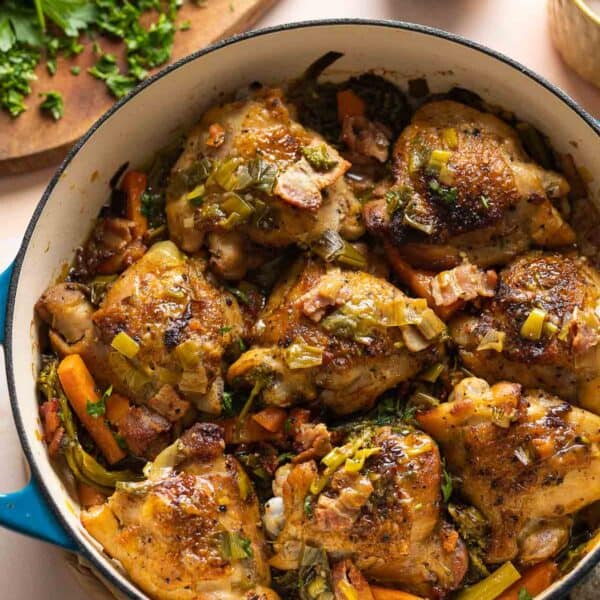 A ceramic casserole dish full of white wine braised chicken thighs and assorted vegetables including carrots and broccoli with fresh parsley chopped on the side.