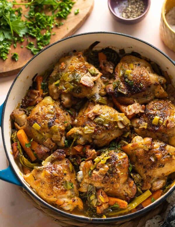 A ceramic casserole dish full of white wine braised chicken thighs and assorted vegetables including carrots and broccoli with fresh parsley chopped on the side.