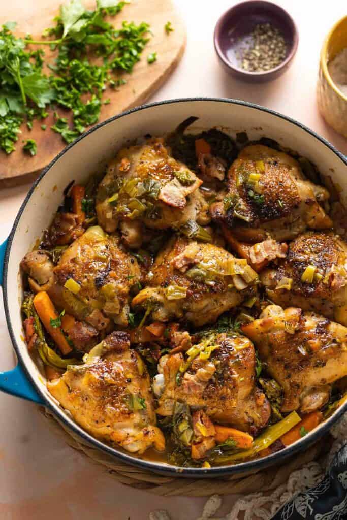 A ceramic casserole dish full of white wine braised chicken thighs and assorted vegetables including carrots and broccoli with fresh parsley chopped on the side.