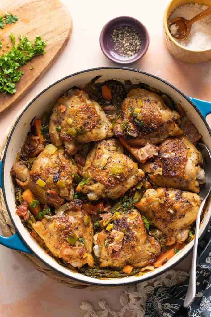 A ceramic casserole dish full of white wine braised chicken thighs and assorted vegetables including carrots and broccoli. A cutting board with fresh chopped parsley to garnish the dish and a serving spoon on the side.