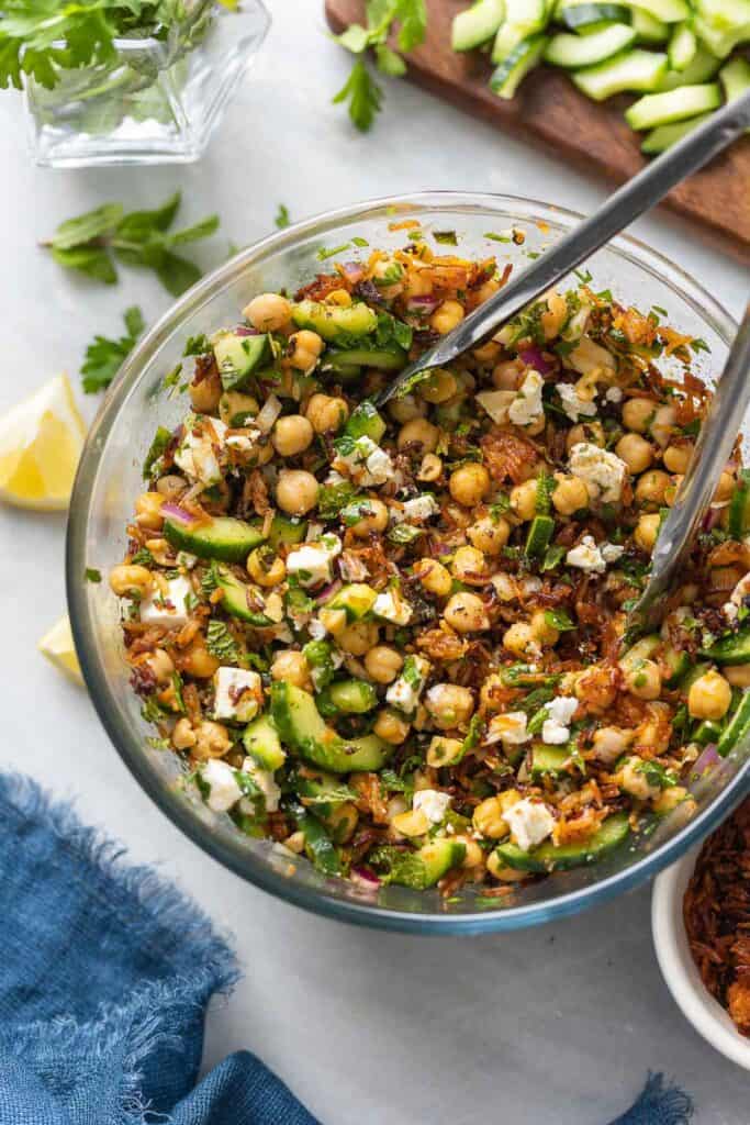 A large bowl full of a Crispy Rice Salad with Lemony Chickpeas & Cucumber and crumbled feta mixed with tongs.