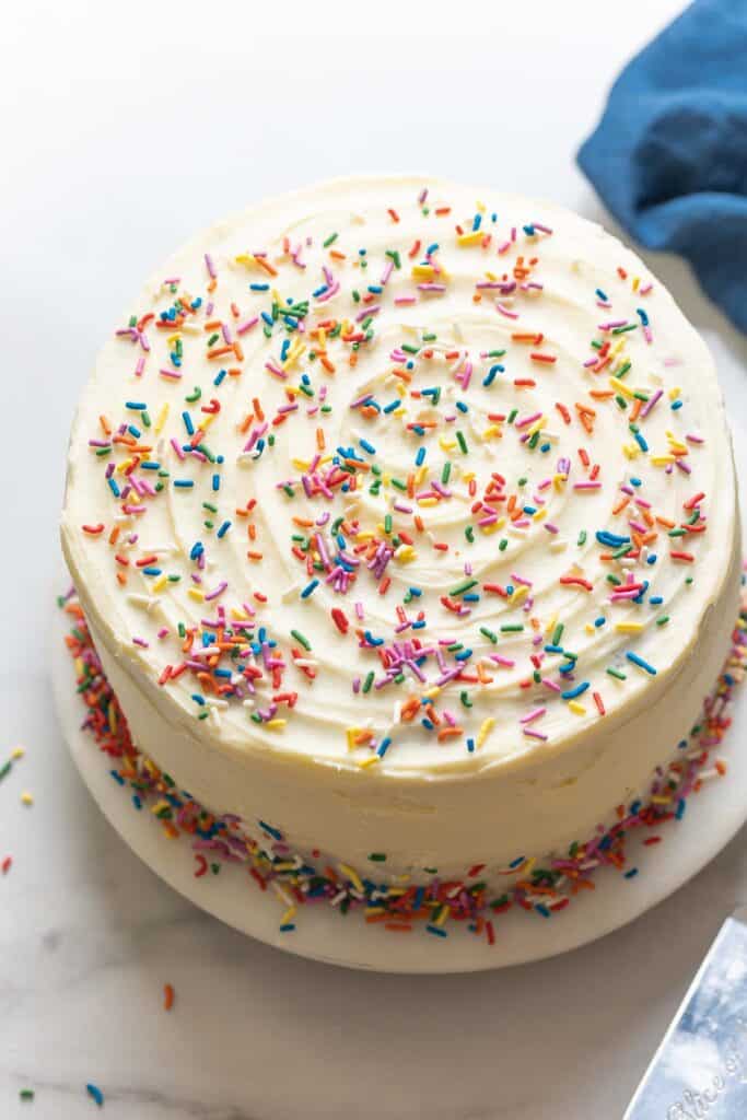 A gluten free confetti cake decorated with vanilla frosting and rainbow sprinkles (AKA 'Jimmies) on a cake platter.