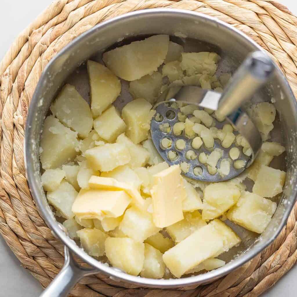 A medium pot full of soft, boiled potatoes with a few pieces of butter and a potato masher.