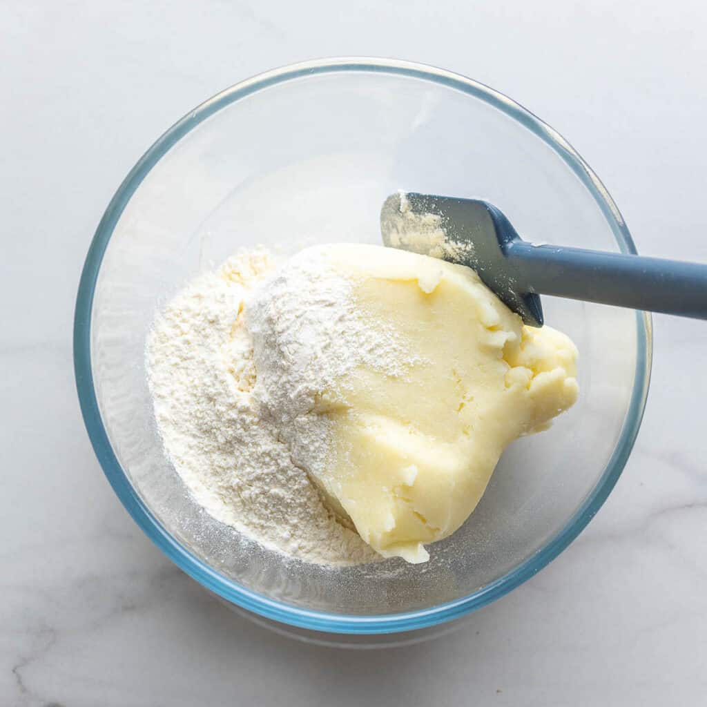 Mashed potato in a mixing bowl with flour, baking powder and salt and a silicone spatula to mix it all together.