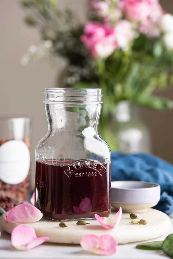 A Kilner storage jar filled halfway full of a a homemade cardamom rose simple syrup with fresh rose petals and cardamom pods scattered around the jar.