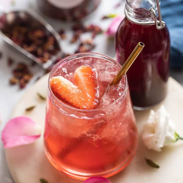 A cardamom rose mocktail in an low ball glass with a heart cut in the shape of a strawberry; rose petals and cardamom pods surround the glass