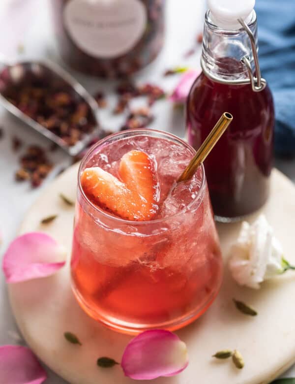 A cardamom rose mocktail in an low ball glass with a heart cut in the shape of a strawberry; rose petals and cardamom pods surround the glass